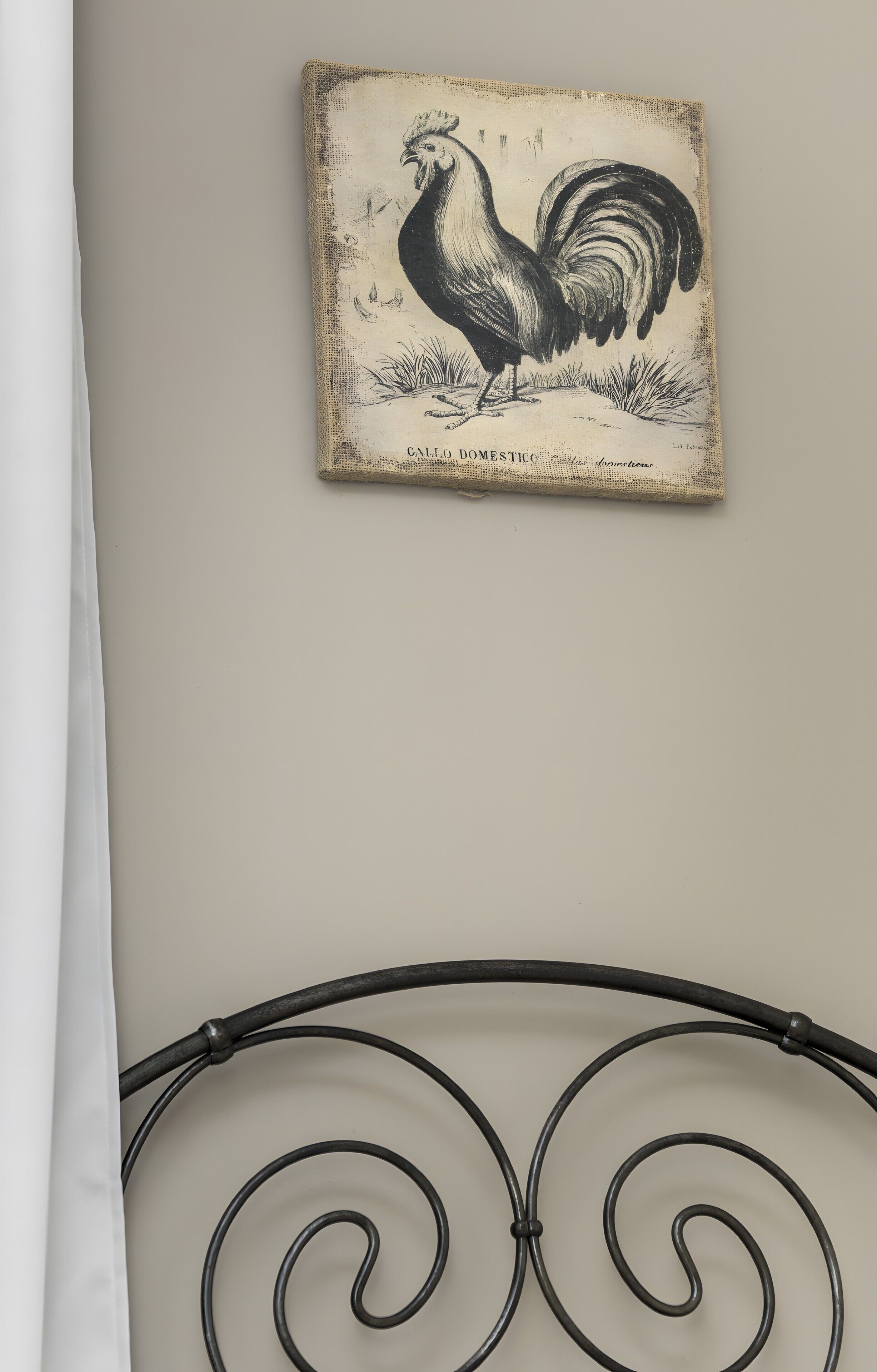 Een aan de muur bevestigd kunstwerk van een haan, een metalen bedframe en een witte gordijn.