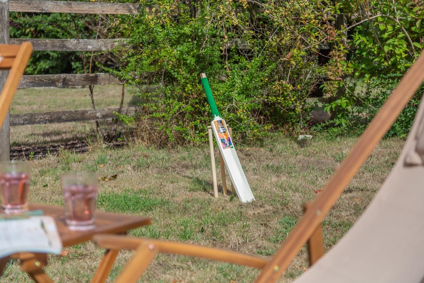 Un bastone da cricket appoggiato a un recinto di legno in una zona erbosa, con fitta vegetazione sullo sfondo.