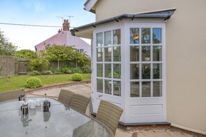 A patio with a glass table and chairs, a white door, and a garden with a hedge and a house in the background.
