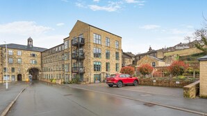 A multi-story building with a central courtyard, a red car parked in the driveway, and a stone wall with flowering plants. - 2 Bed in Haworth (oc-c37865) (Haworth)