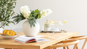 A wooden table with a white vase of flowers, a bottle of PENTIRE, two glasses of white wine, a bowl of cantaloupe, and an open magazine. - 4 Bed in Rock (oc-l50184) (Rock)