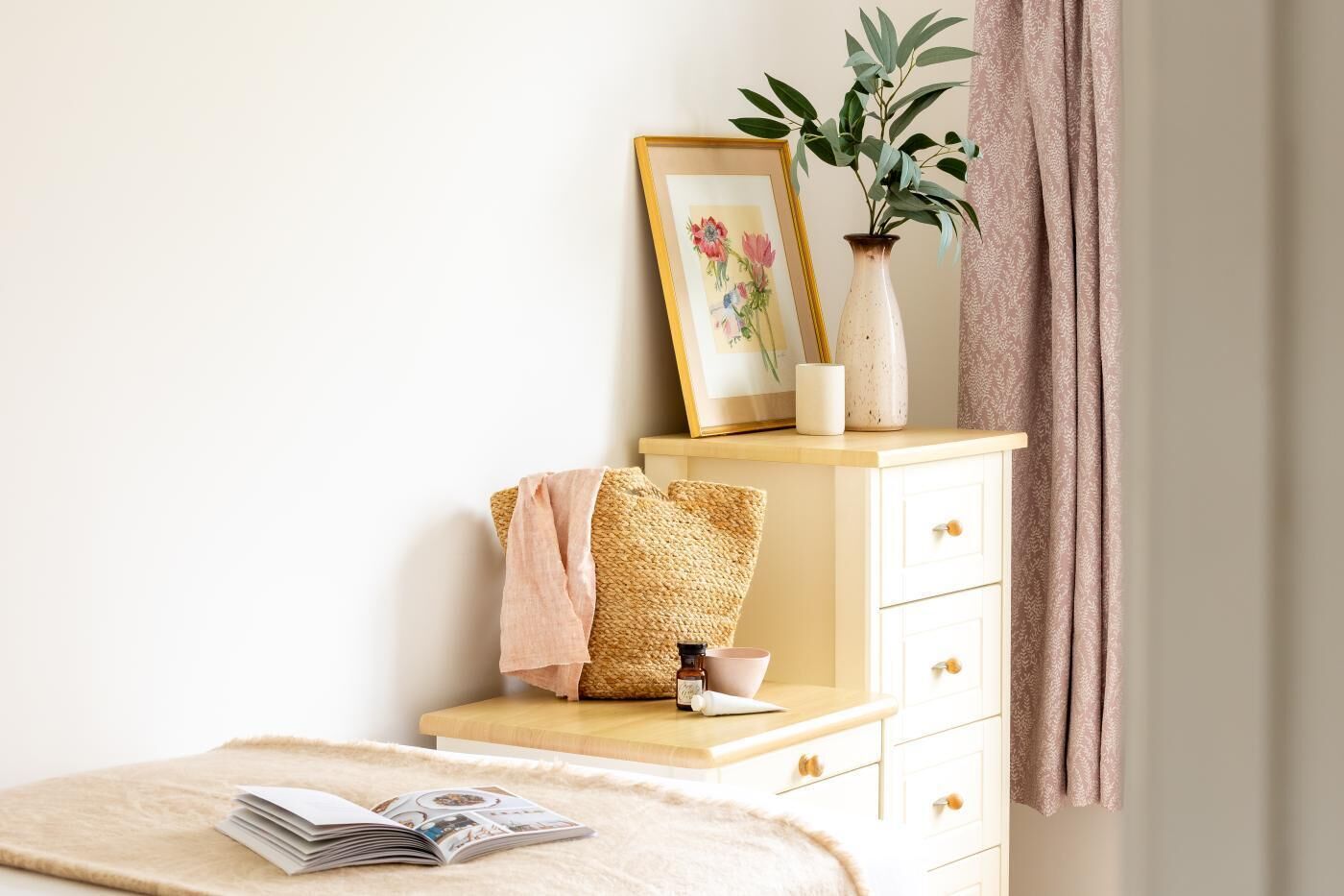 A neatly arranged bedroom with a bed, a bedside table with a book, a vase with a plant, and a framed picture on the wall.