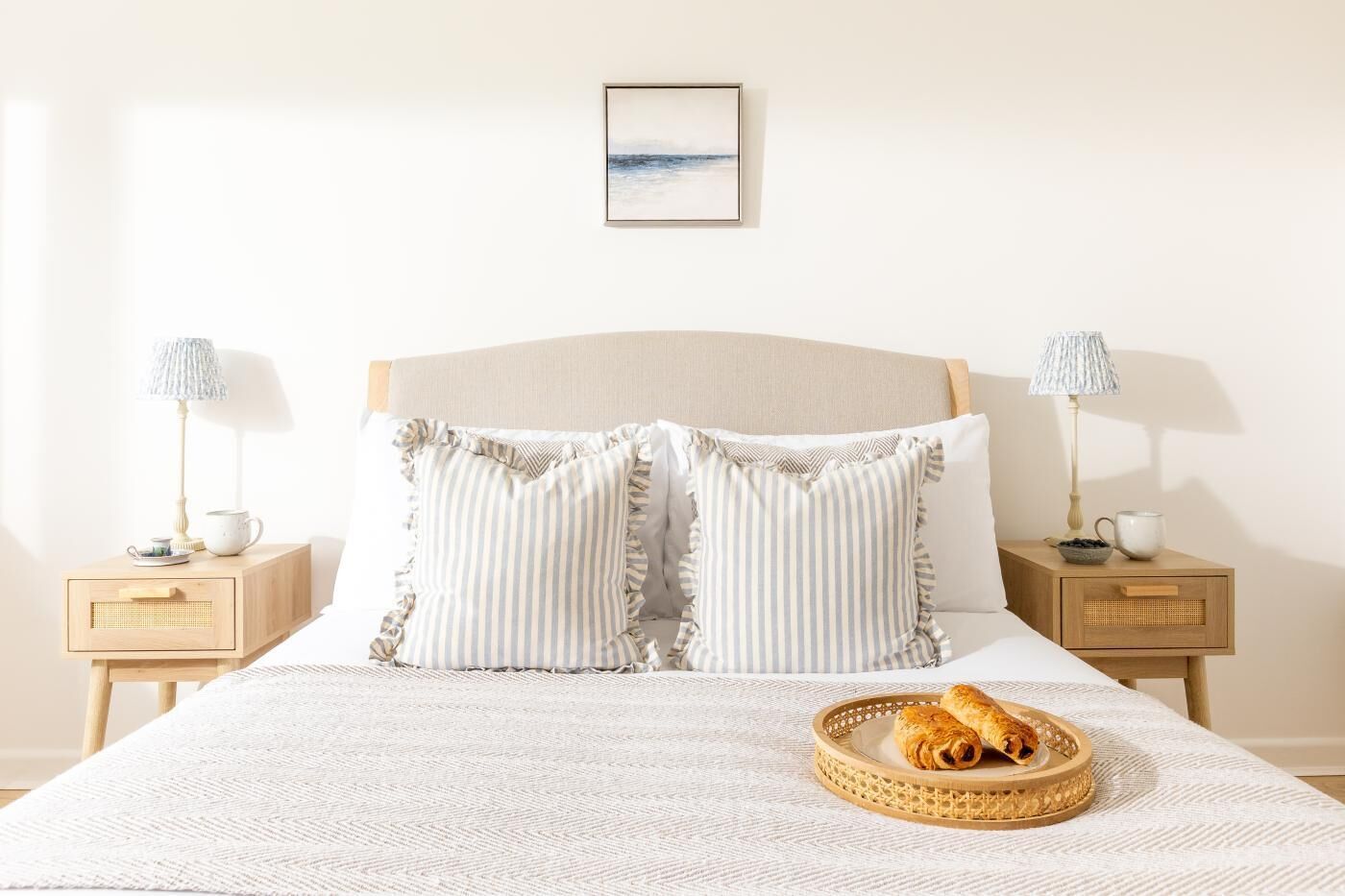 Een netjes opgemaakt bed met gestreepte kussens, een nachtkastje met een lamp en een dienblad met gebak.