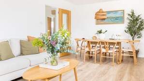Ein modernes Wohnzimmer mit einer Couch, einem Esstisch mit Stühlen, einer Vase mit Blumen, einem Gemälde an der Wand und einem Holzboden.
