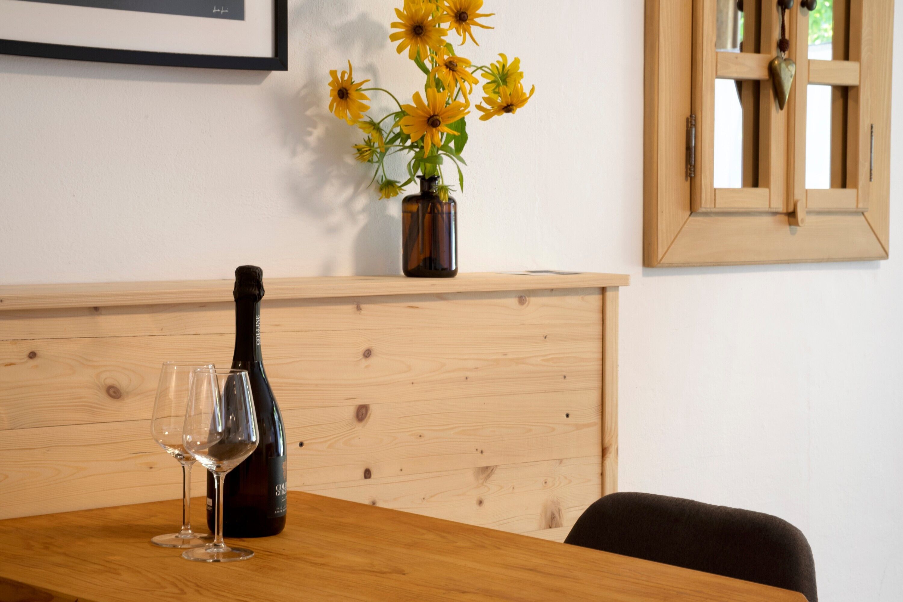 A wooden table with a wine bottle and two glasses, a vase with yellow flowers, and a framed picture on the wall.