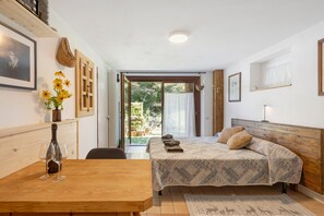 A bedroom with a bed, a wooden table with wine glasses, a vase with flowers, and a view of the outside.