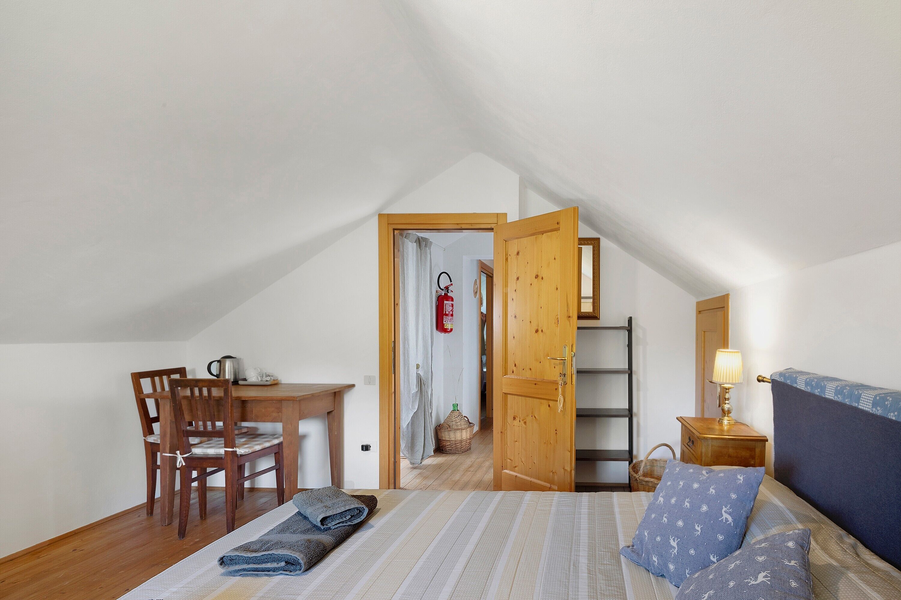 A bedroom with a bed, a desk, a chair, a nightstand, and a wooden door leading to another room.