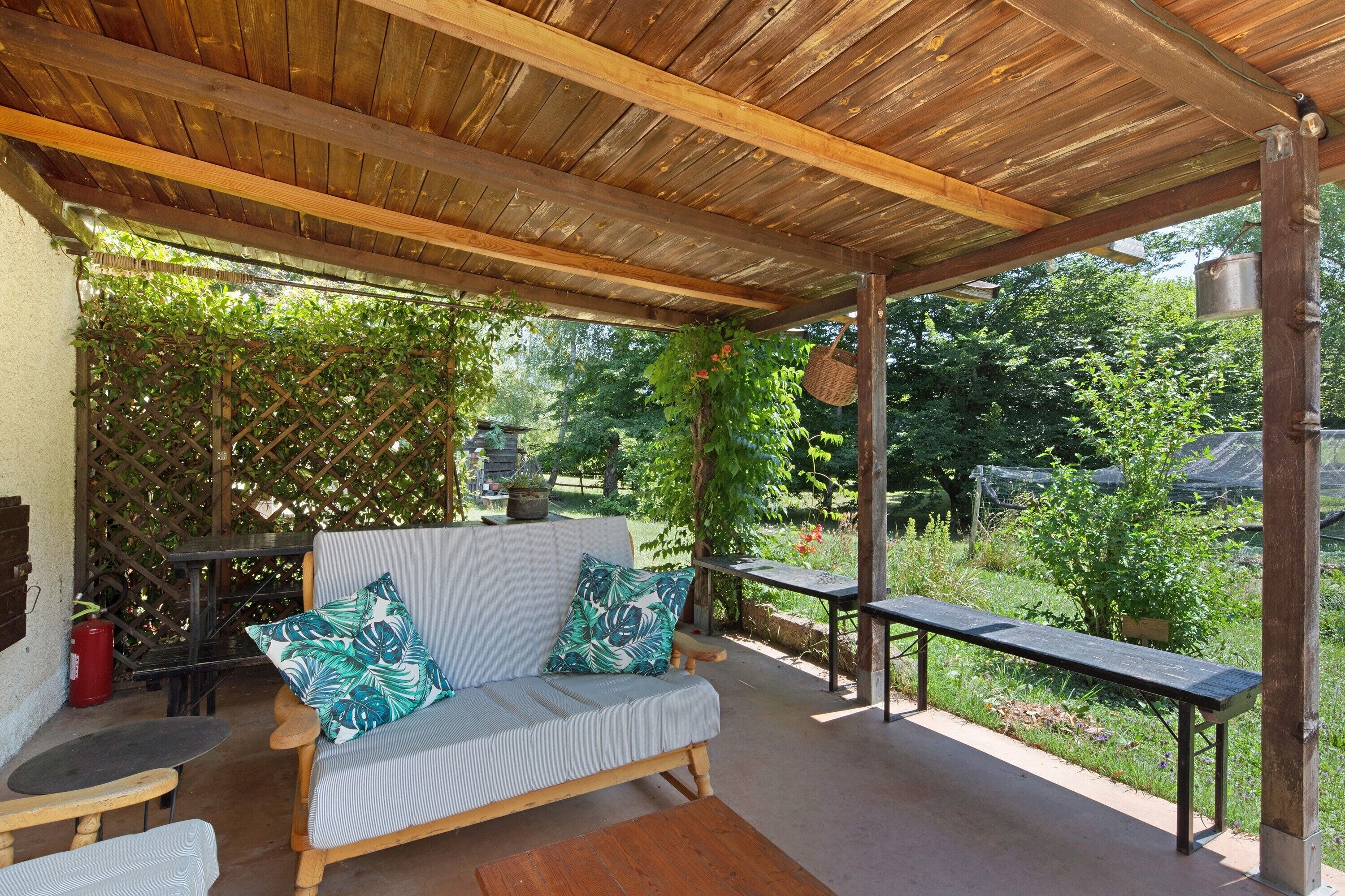 En udendørs terrasse med et træloft, en polstret sofa med mønstrede hynder, en bænk og et bord.