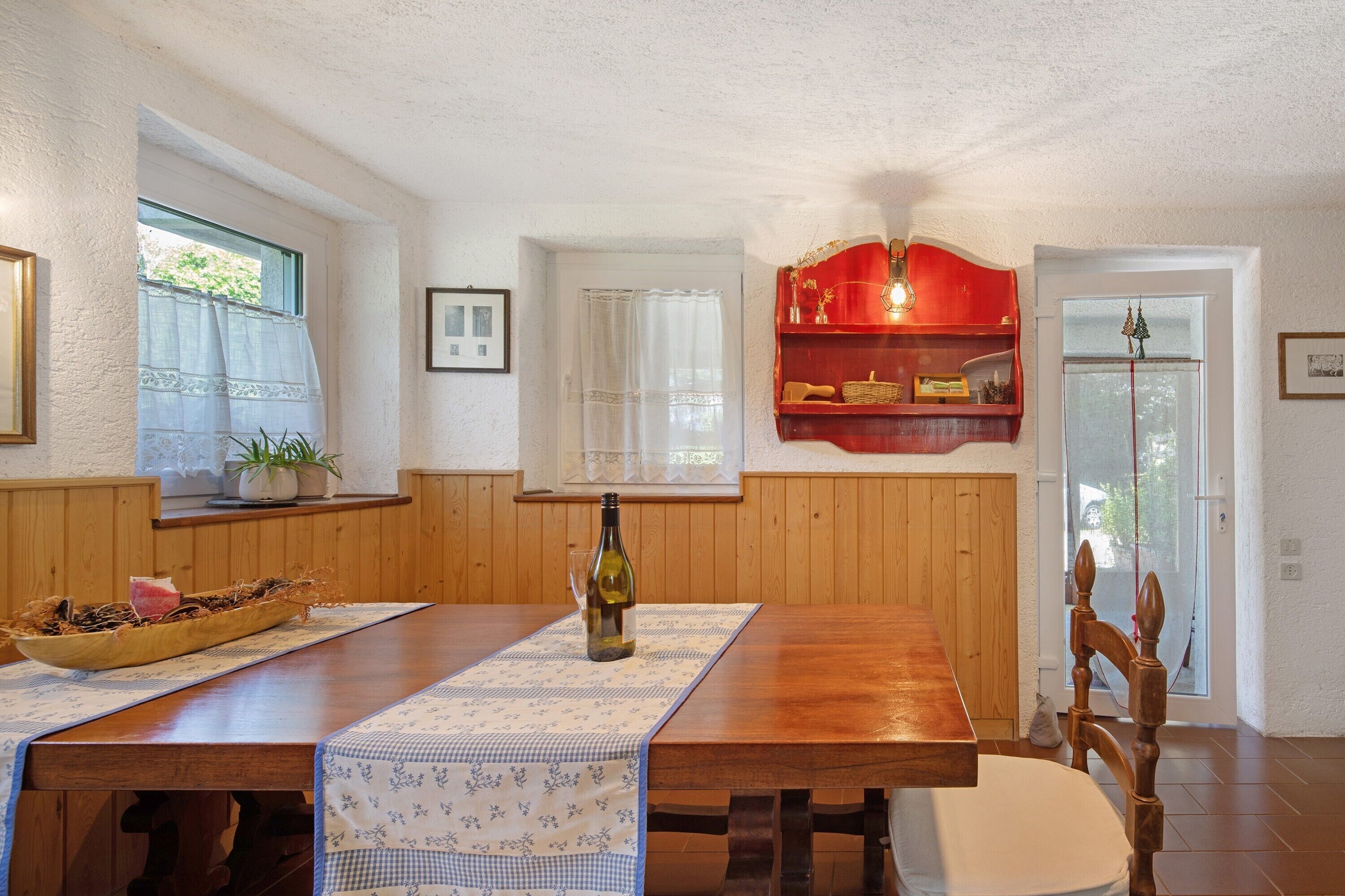 A dining area with a wooden table, chairs, a bottle of wine, and a red shelf with decorative items.