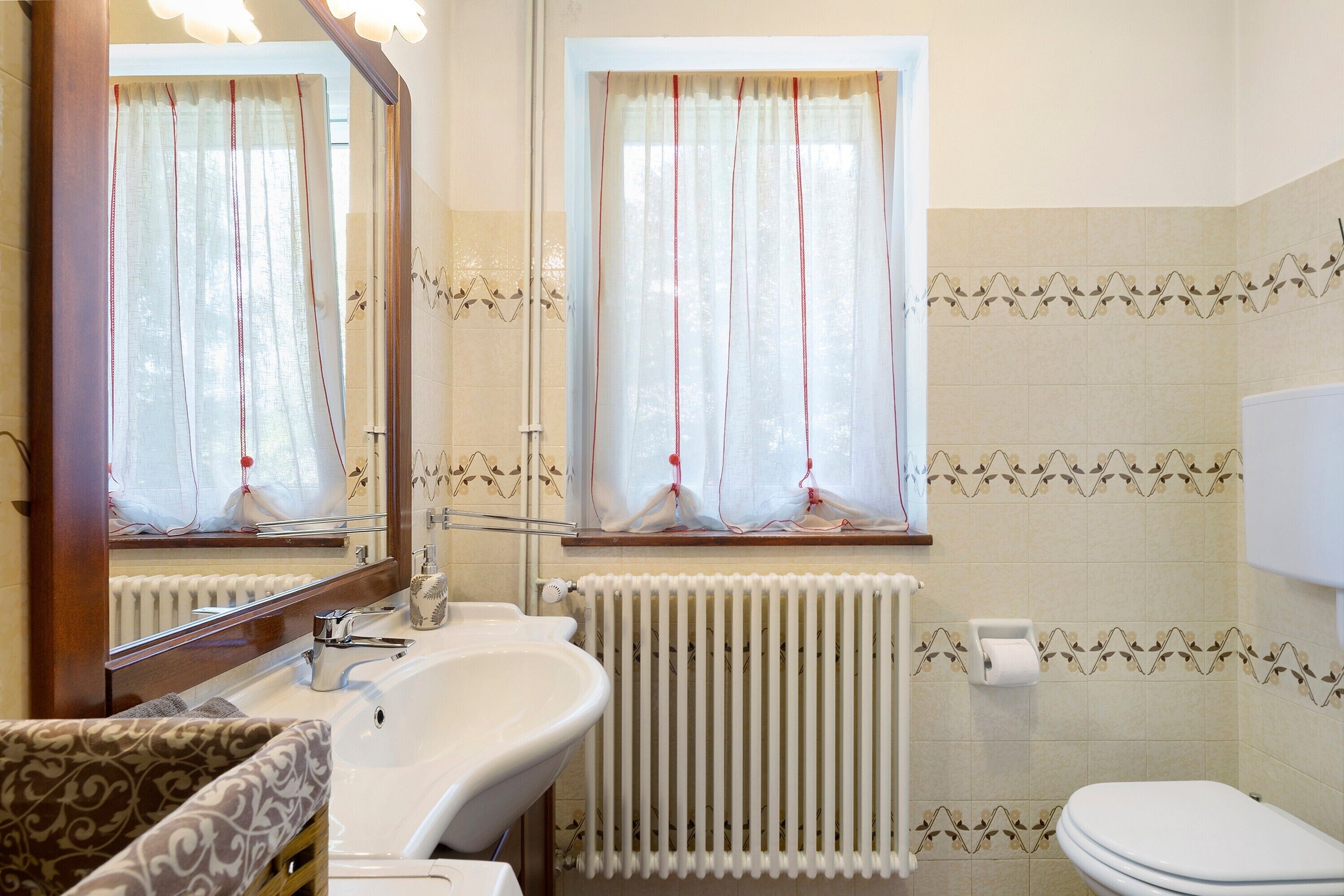 A bathroom with a sink, mirror, window with curtains, radiator, and toilet.