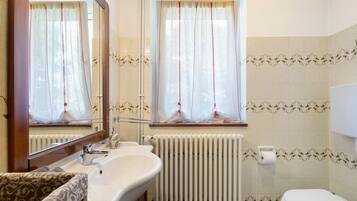 A bathroom with a sink, mirror, window with curtains, radiator, and toilet.
