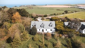 Een groot wit huis met een grijs dak, omringd door bomen en velden, met uitzicht op de zee op de achtergrond.