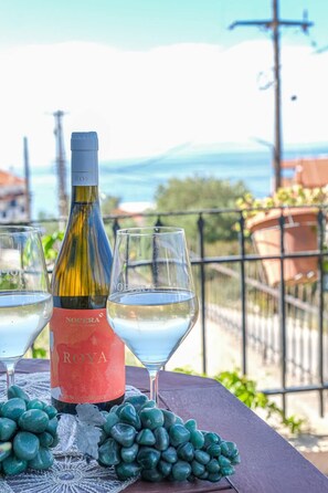 A bottle of Notera ROYA wine, two glasses, and a bunch of grapes on a table with a view of the sea and buildings. - Breeze - Getaway in Samos w Serene Sea View (Samos)