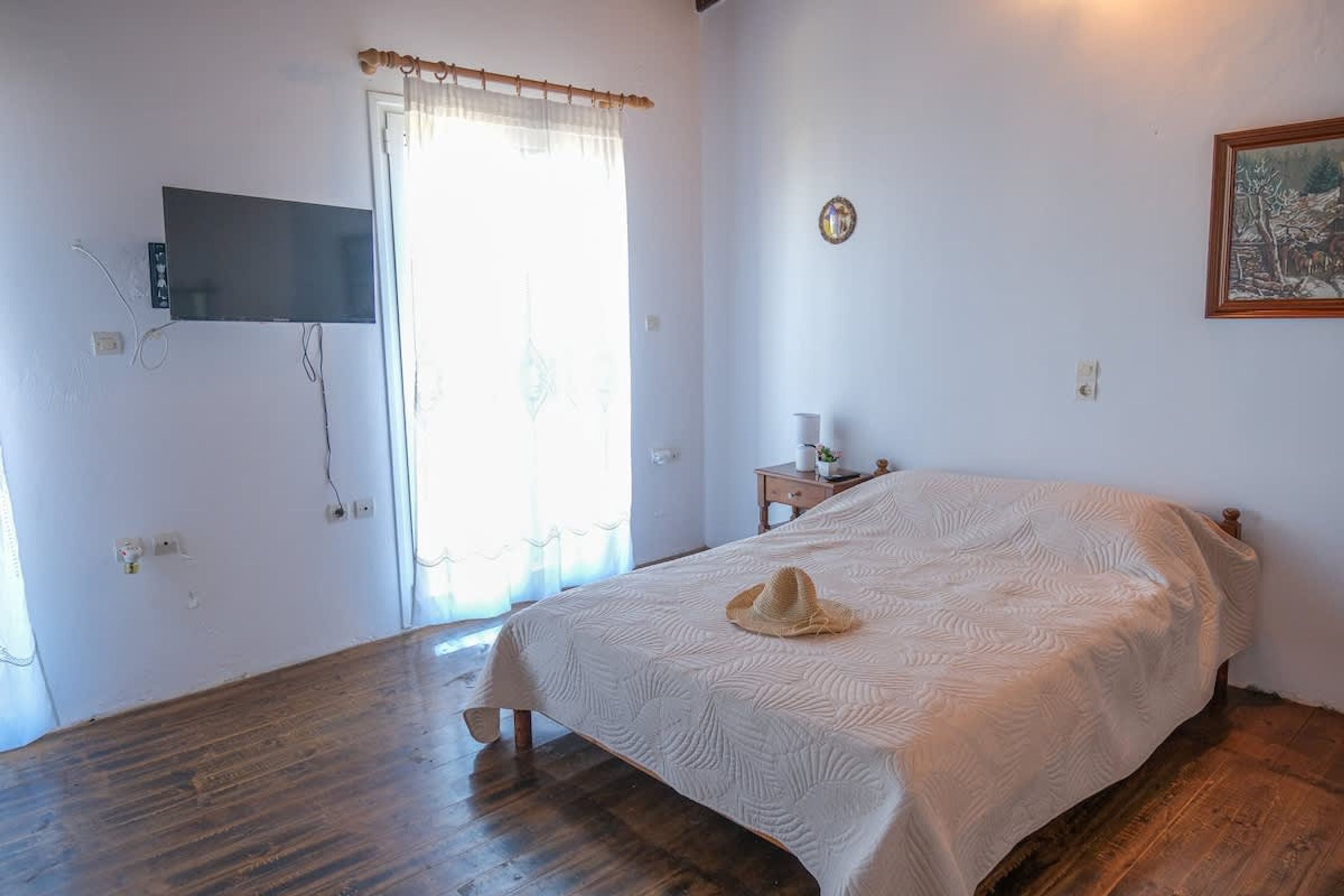 A bedroom with a bed, a television mounted on the wall, a window with curtains, a small bedside table, and a wall clock.