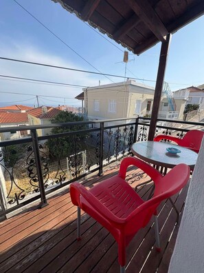 A balcony with a wooden floor, a round table with chairs, and a view of buildings in the background. - Little Nest in Koumeika Samou (Samos)