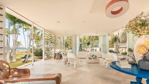 A spacious living area with a large window overlooking palm trees and a pool, a white sofa, a coffee table, and a blue accent wall. - So Exclusive Beachfront Villa at Las Terrenas (Las Terrenas)