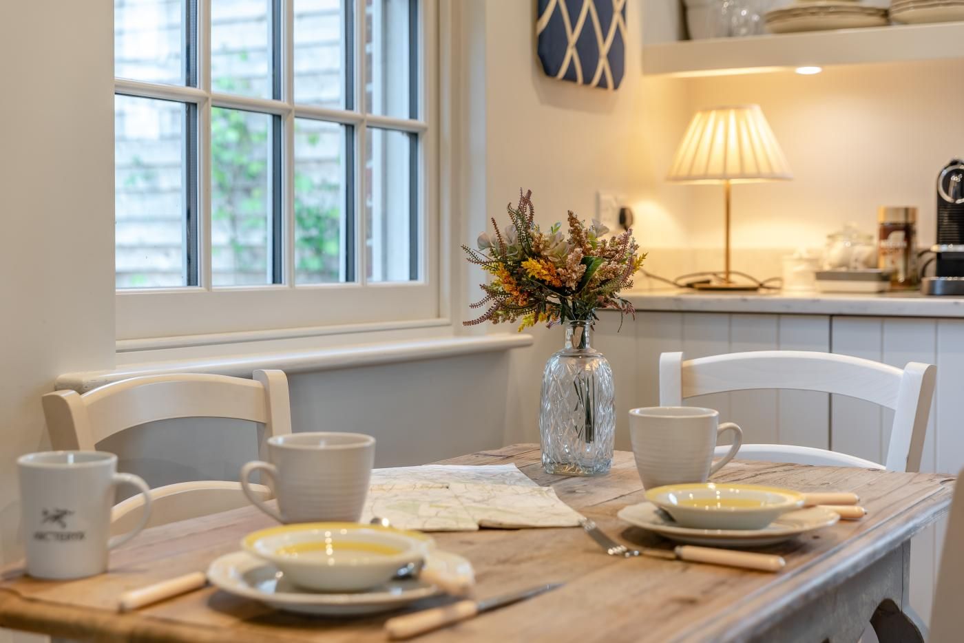 A dining table with chairs, a vase with flowers, and a lamp in a room with a window and a kitchen in the background.