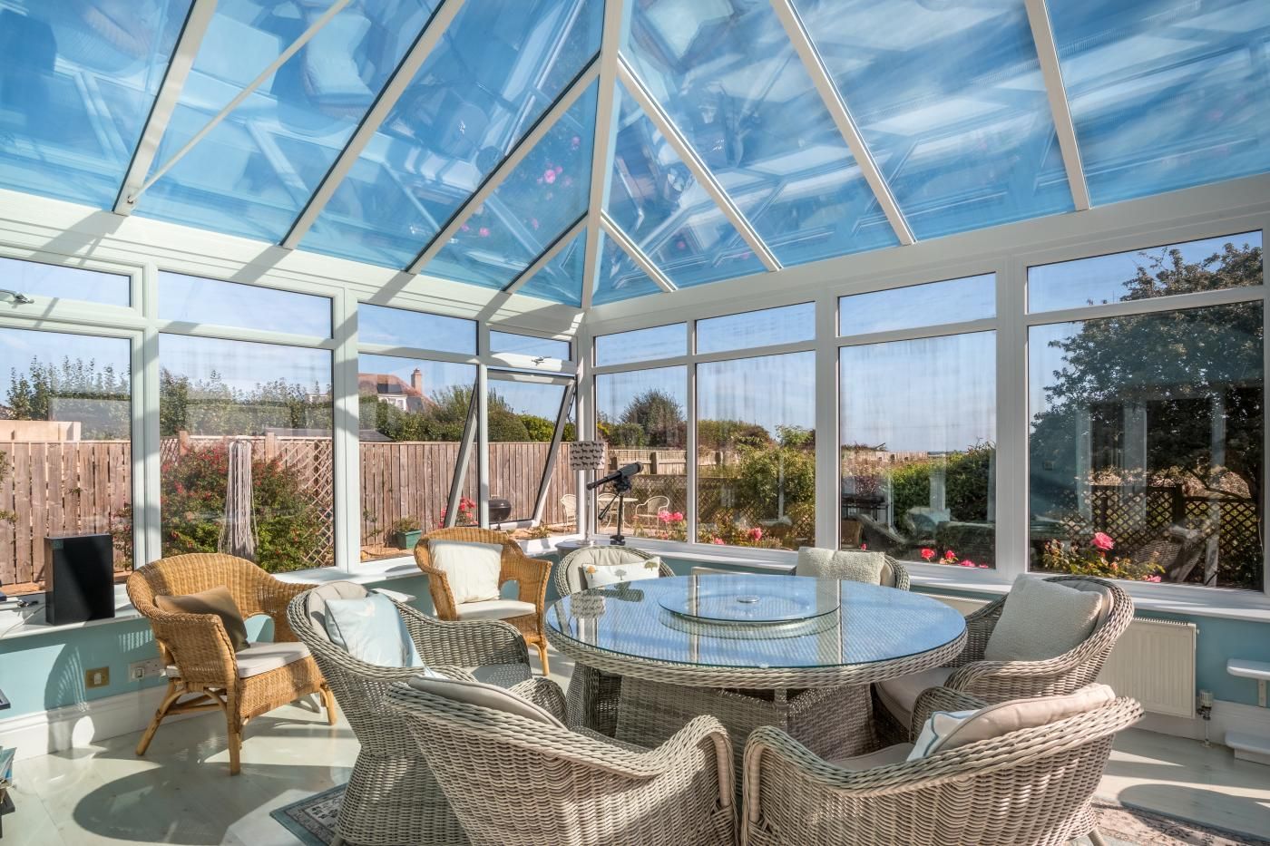 A conservatory with a glass ceiling, wicker furniture, and a view of a garden.