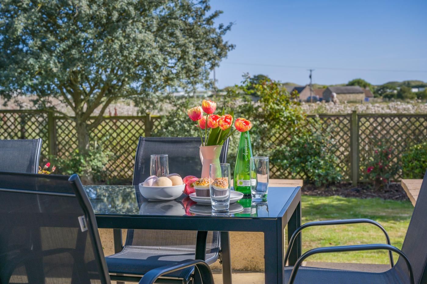 Une table à manger extérieure avec des chaises, un saladier rempli de fruits, un vase avec des fleurs et des bouteilles d’eau.
