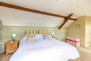 A bedroom with a large bed, wooden headboard, two bedside tables with lamps, a small bedside table with a book, and a red and white patterned ottoman. - 2 Bed in Dulverton (oc-h34793) (Brushford)