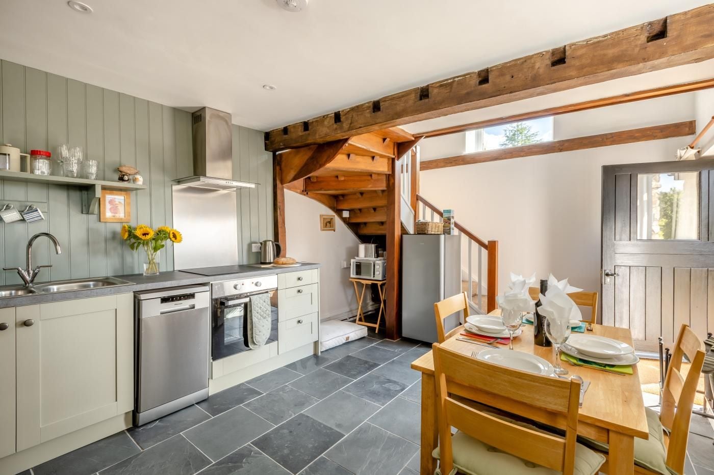 A kitchen with a dining area, featuring a wooden staircase, a sink, and a dining table set with plates and cutlery.