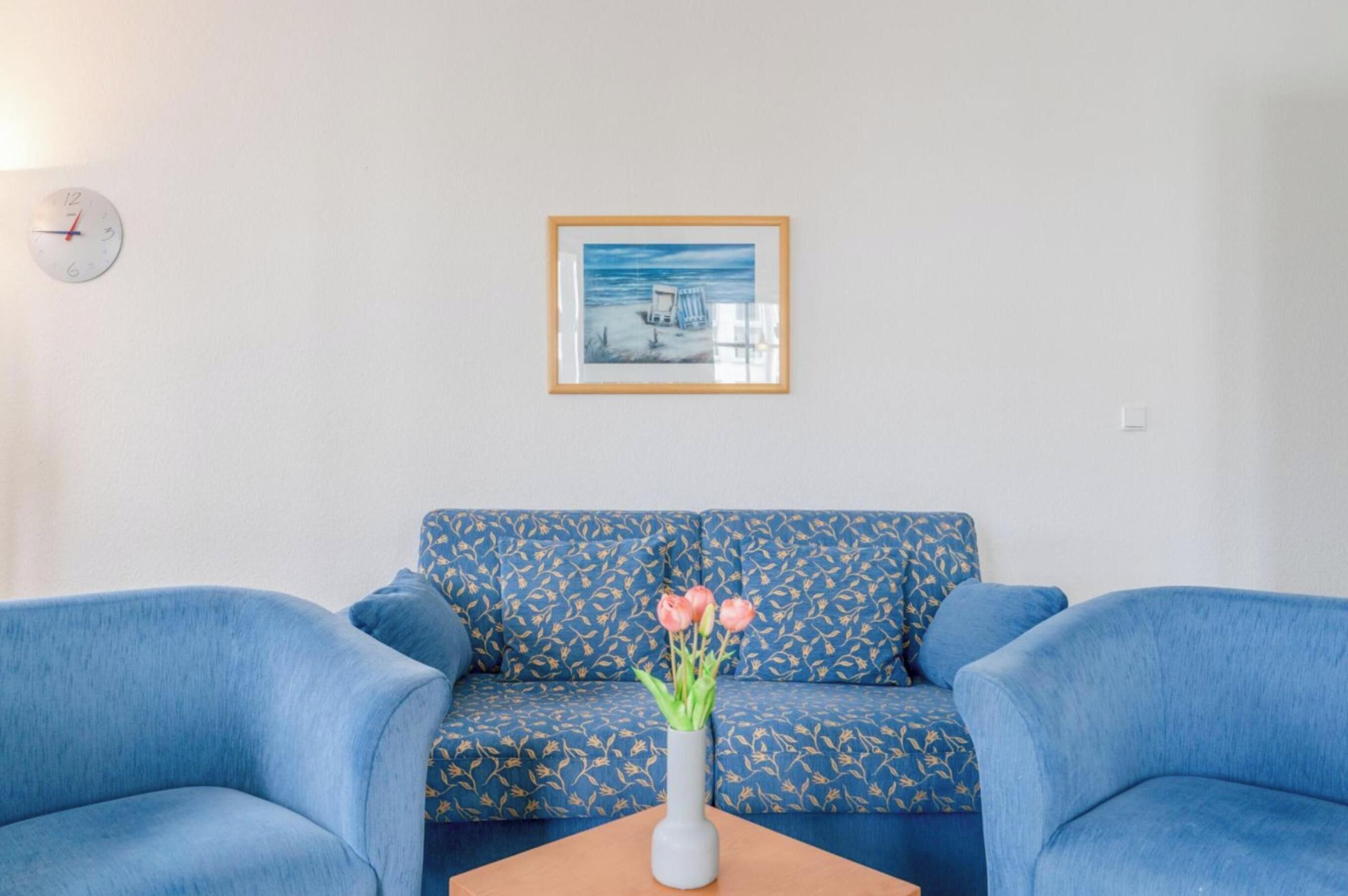 A room with a blue sofa, a small table with a vase of flowers, a framed picture on the wall, and a clock.