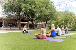 Eine Gruppe von Menschen sitzt auf Yogamatten im Freien, mit Bäumen und einem Gebäude im Hintergrund.
