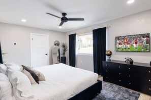 Ein Schlafzimmer mit Bett, Deckenventilator, Schminktisch, Spiegel, Fenster mit Vorhängen und einem Foto einer Sportmannschaft an der Wand.