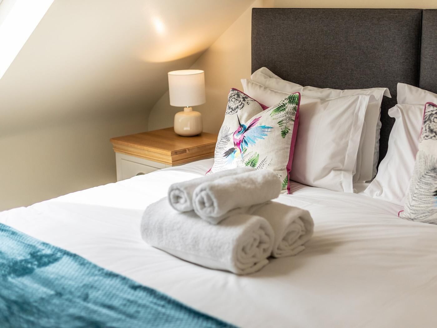 Una cama bien hecha con toallas enrolladas, una lámpara de noche y almohadones decorativos.