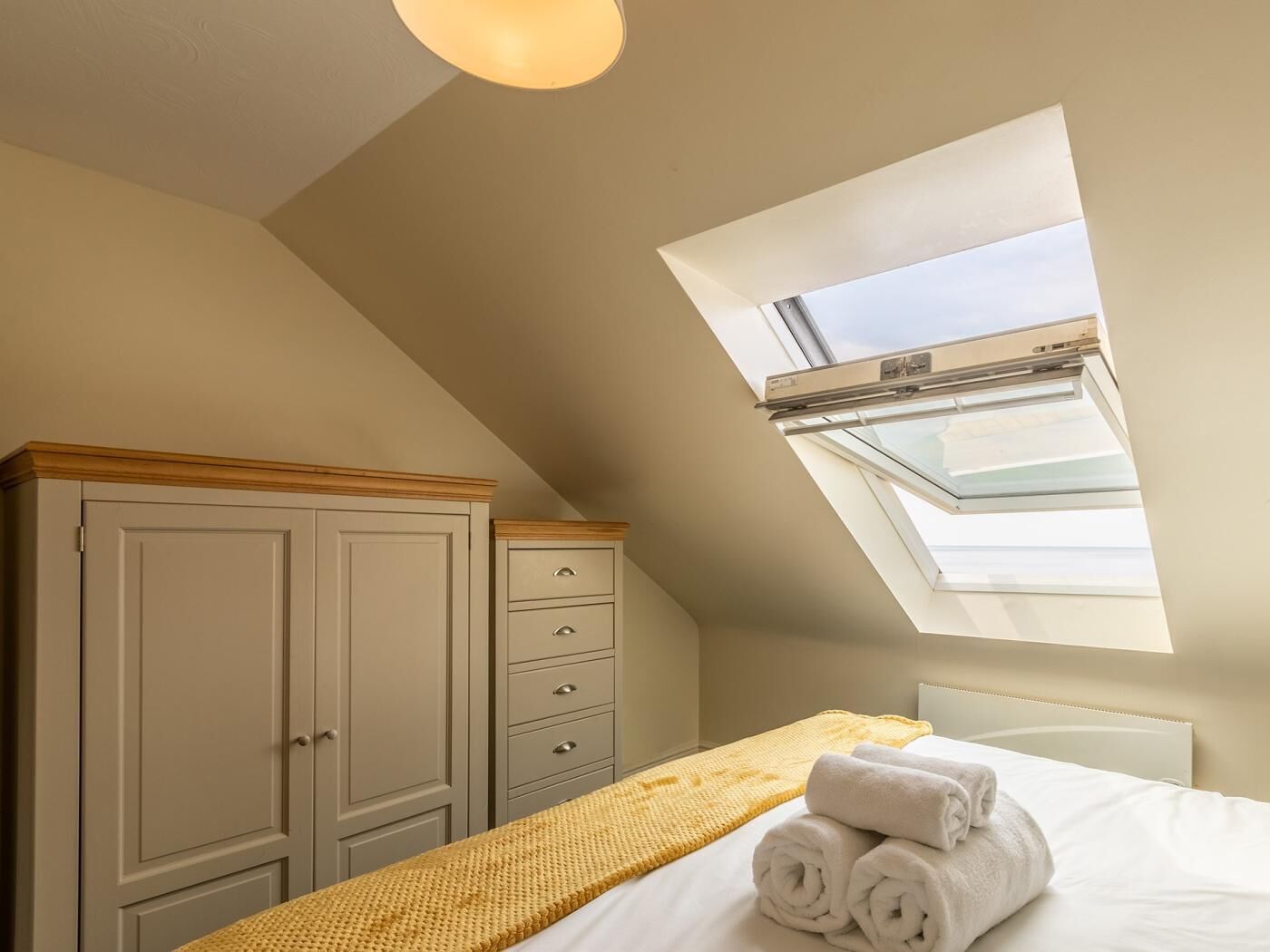 Ein Schlafzimmer mit Dachfenster, einem Bett mit aufgerollten Handtüchern, einer Holzkommode und einer gelben Decke.