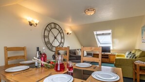 A dining area with a wooden table set for four, a clock on the wall, and a living area with a sofa and television. - 3 Bed in Braunton (oc-87112) (Croyde)