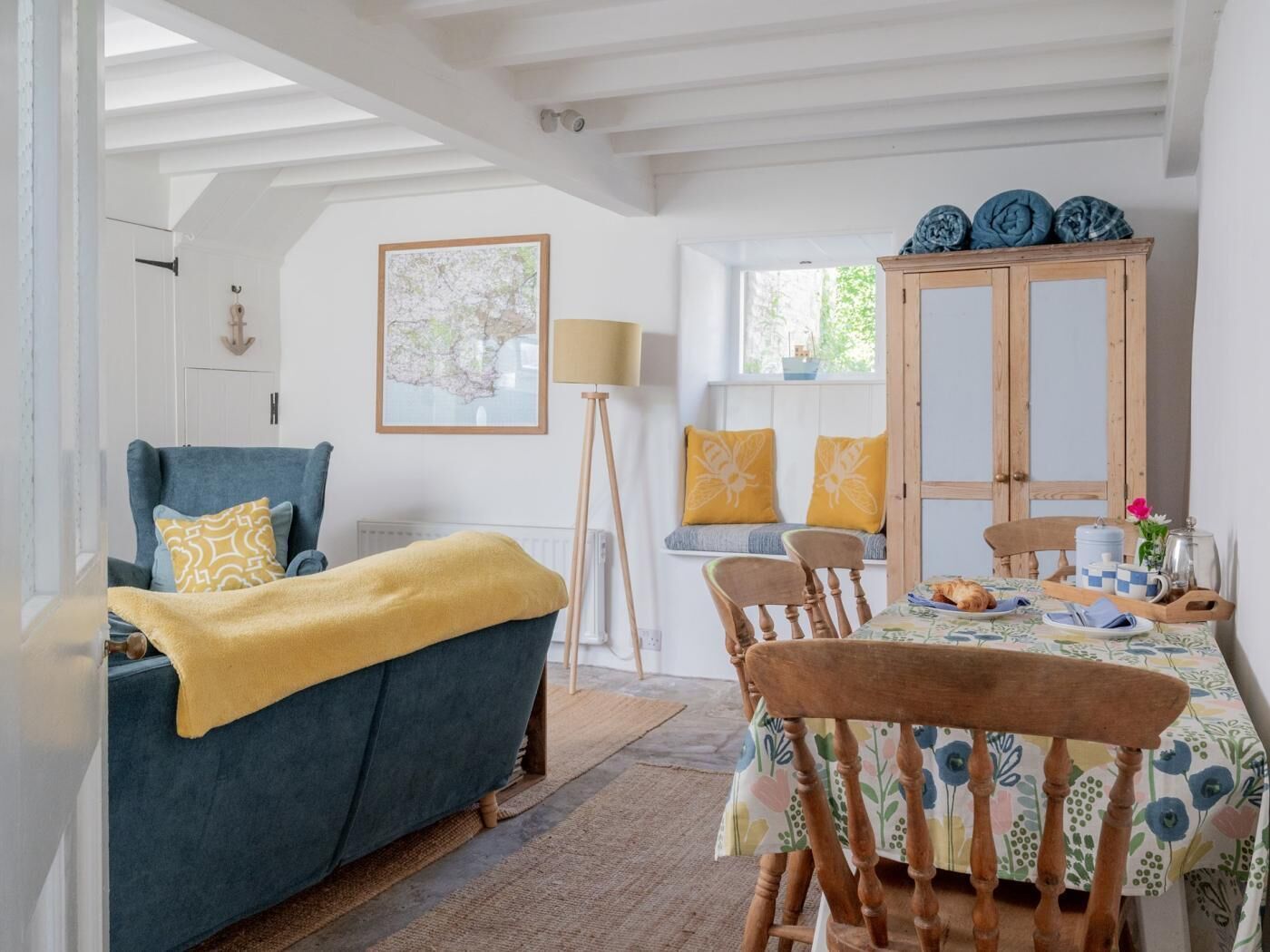Une pièce chaleureuse avec un canapé bleu, des chaises en bois, une table à manger recouverte d’une nappe à motifs floraux et un buffet contenant de la vaisselle bleue.