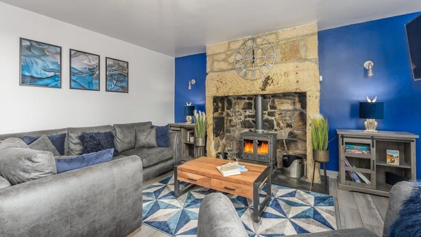 A living room with a stone fireplace, grey sofas, a coffee table, and blue walls. - 2 Bed in Warkworth (oc-coq103) (Warkworth)