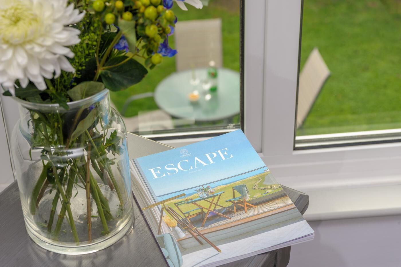 A clear glass vase with flowers and a book titled 'ESCAPE' on a table by a window.