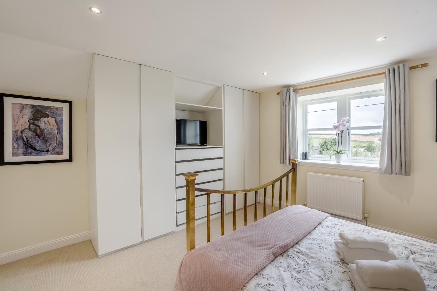 Een slaapkamer met een bed, een kledingkast, een televisie, een raam met gordijnen en een schilderij aan de muur.