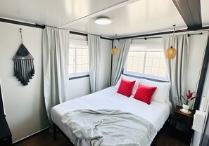 Una cama con ropa de cama blanca y almohadas rojas, un adorno de macramé colgante y una mesita de noche con un florero.