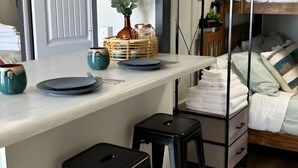 Una cocina moderna con encimera de tonos claros, dos taburetes negros, un aparador de madera con toallas apiladas y una cama con ropa de cama blanca al fondo.
