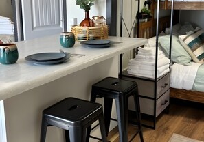 Una cocina moderna con encimera de tonos claros, dos taburetes negros, un aparador de madera con toallas apiladas y una cama con ropa de cama blanca al fondo.