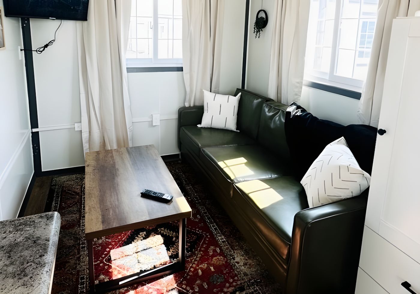 Una sala de estar con un sofá, una mesa de centro, un televisor montado en la pared y un ventanal con cortinas.