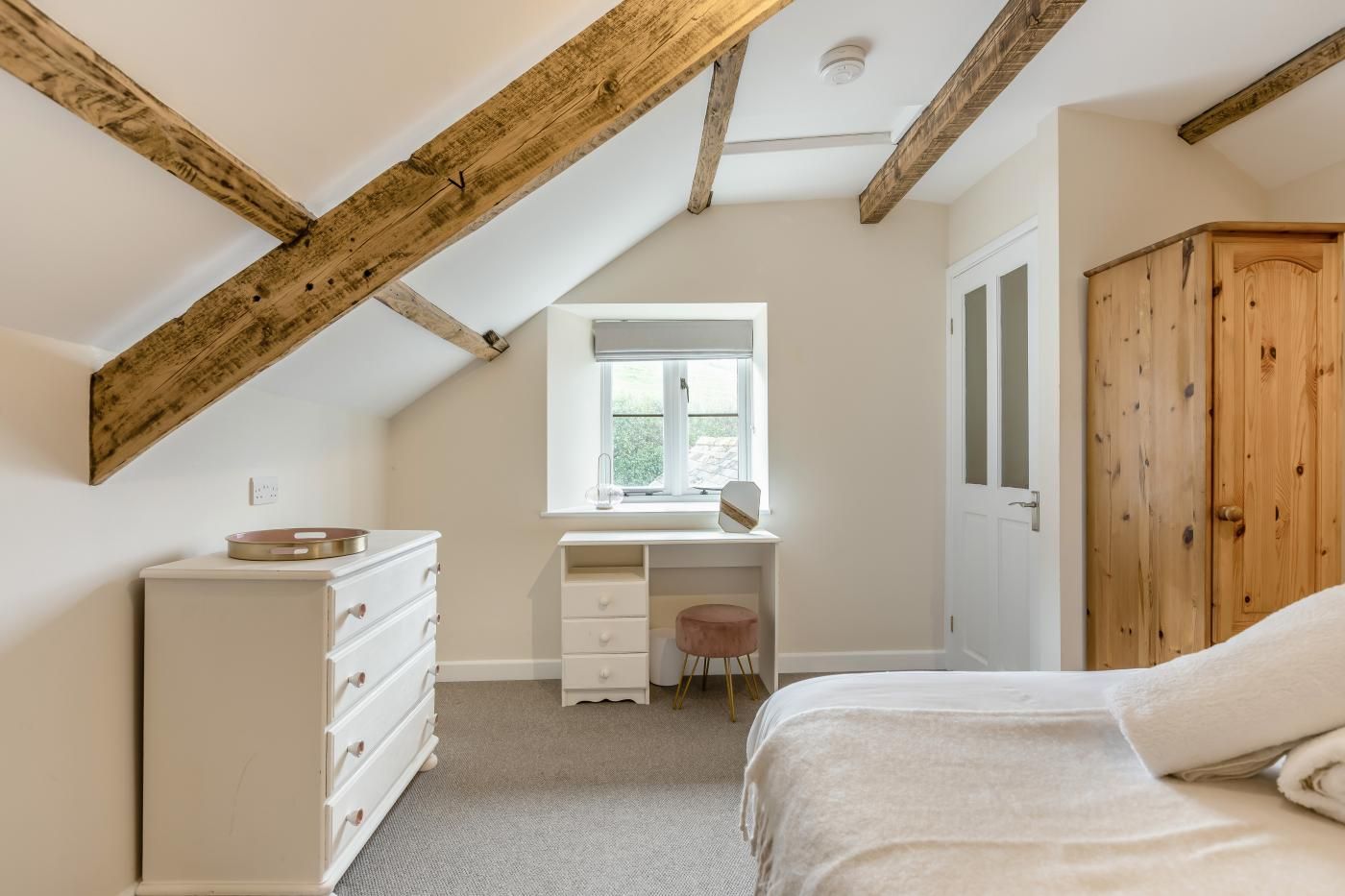 A bedroom with a sloped ceiling, a wooden wardrobe, a bed, a dresser, a vanity with a mirror, and a window.