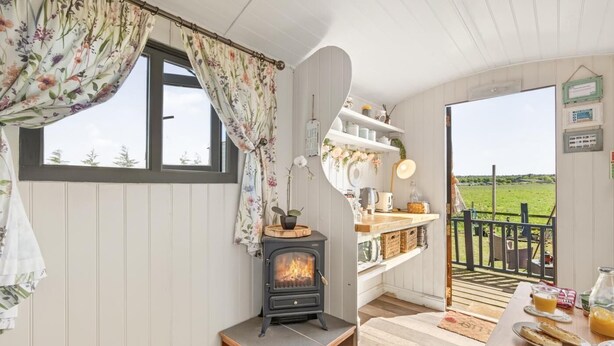 A cozy room with a fireplace, a window with floral curtains, a wooden bench, and a view of a green field through an open door.