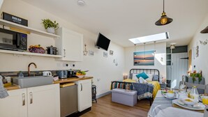 Une cuisine compacte équipée d’un four à micro-ondes, d’un évier et de divers appareils électroménagers. On y trouve un lit avec vue sur le coucher de soleil, une table à manger avec des articles pour le petit-déjeuner et un téléviseur fixé au mur.