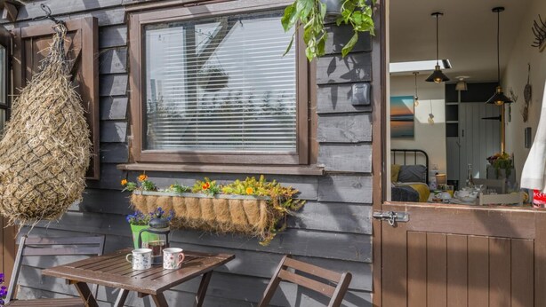 Ein Holzfassade mit Fenster, einer hängenden Blumenampel, einem kleinen Tisch mit Stühlen und einem Pflanzkasten mit Blumen.