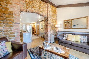 Un salon chaleureux avec une arche en pierre, un canapé agrémenté de coussins décoratifs, une table basse avec des livres et des télécommandes, et un grand miroir au mur.
