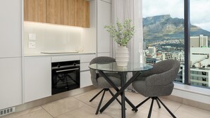 Una cuina moderna amb una taula de vidre i caires, un armari de fusta i vista a un paisatge urbà i muntanyes.