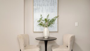 Une pièce avec une table ronde, deux chaises, un vase avec des plantes vertes et un grand tableau abstrait accroché au mur.