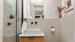 Un baño con ducha, lavamanos, inodoro, espejo y un florero con flores.