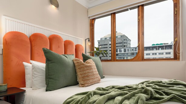 Una cama con una manta verde, almohadas naranjas y verdes, y vistas de edificios a través de una ventana.