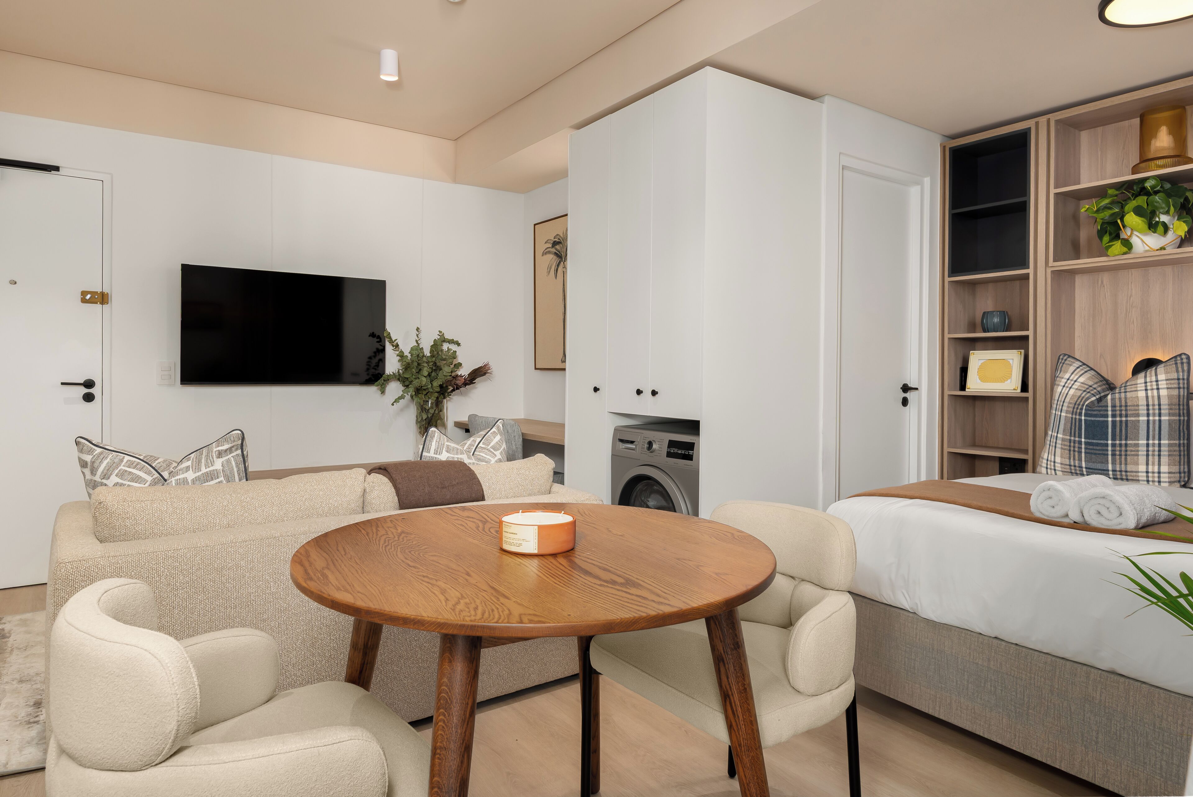 Una sala moderna con un sofá, una mesa redonda de madera, un televisor, una lavadora y una cama con toallas dobladas.
