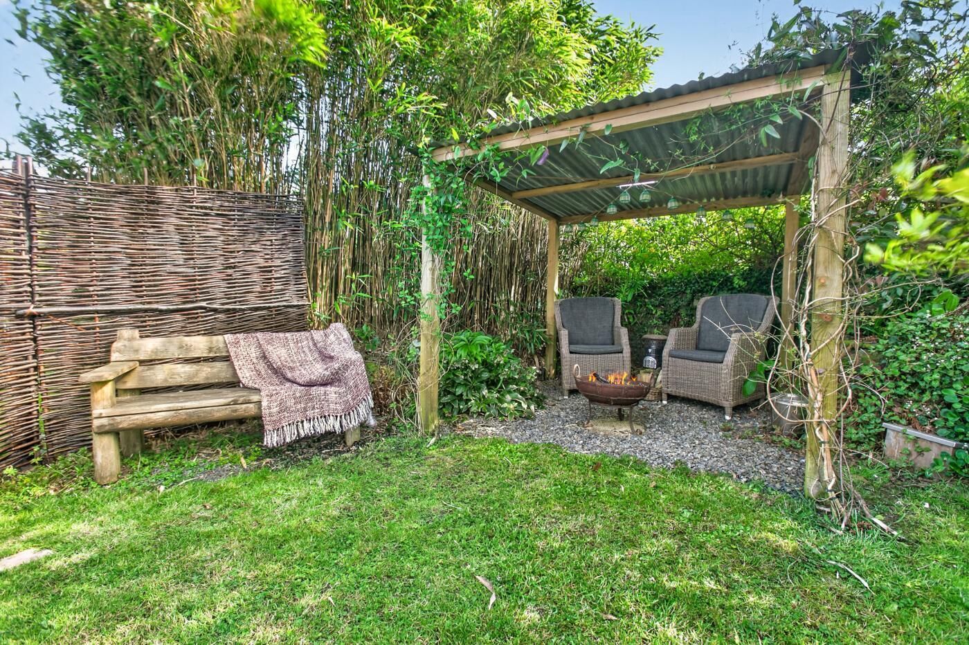 A wooden bench with a blanket, wicker chairs, a fire pit, and a pergola in a garden setting.